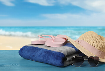 Obraz na płótnie Canvas Beach towel, flip flops, hat and heart shaped sunglasses on light blue wooden surface near seashore