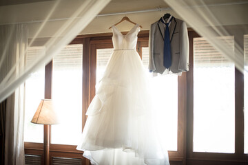 Elegant wedding dress hanging on rack in the room by the window.