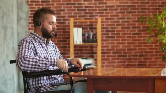 Hipster man in wheelchair finishes working on laptop, closes it, puts it on his laptop. Disabled person wearing headphones with microphone works remotely for people with disabilities