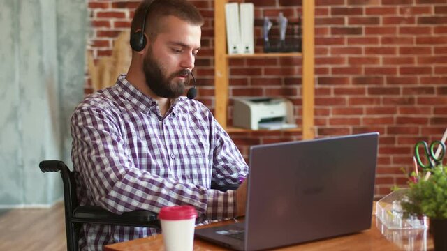 Man is sitting in wheelchair, using laptop with headset, talking via video link and recording important things, side view. Hipster man uses laptop work. Concept of remote work for disabled