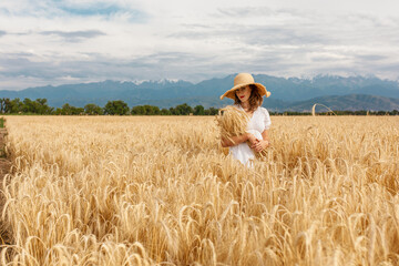 Naklejka na ściany i meble a young attractive woman in a white dress and a straw hat on a field of ripe cereals against a blue sky with clouds in autumn, the concept of harvesting, agribusiness and agriculture