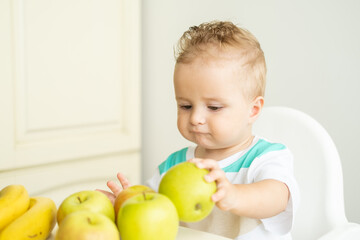 cute baby boy sitting at the table in child chair eating apple on white kitchen.