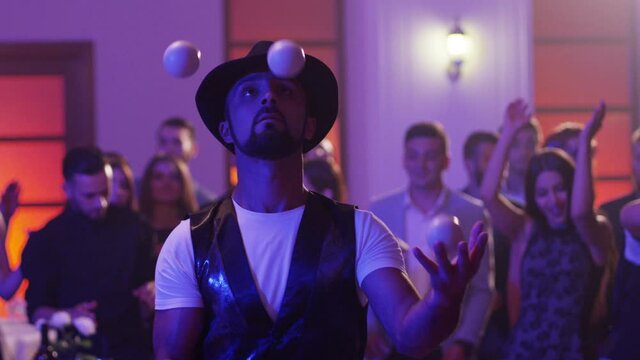 Juggler showing tricks with white balls at colorful party . Man juggling four balls at night party. Many out of focused people in the background . Shot on RED DRAGON Cinema Camera in slow motion.