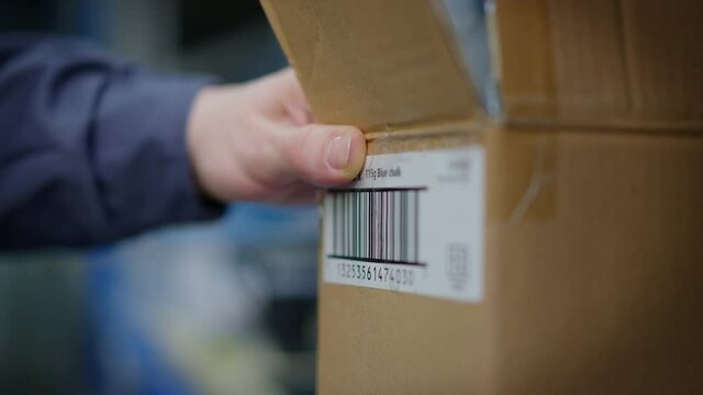 Close-up male hand scanning shipment barcode on cardboard box in terminal warehouse. Unrecognizable Caucasian male inspector checking goods delivery indoors in industrial storage. Modern technologies