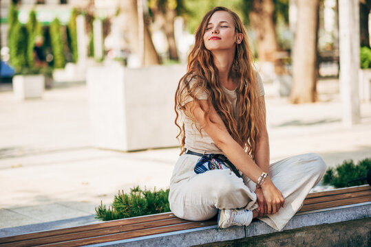 Horizontal image of pretty young woman with long hair resting on the street. Full-length view of female university student sitting outside waiting for her colleague outdoors.