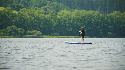Boy rowing on stand up paddle boarding (SUP) paddling along the calm lake. The concept of...