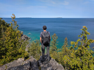 Obraz premium Young man with backpack standing on a high rocky cliff edge overlooking the sea on a sunny day. Bruce Peninsula National Park, Georgian Bay, Ontario, Canada. Hiking, camping, active lifestyle concept.