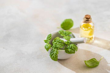 Transparent bottle of basil essential oil, fresh basil leaves in a ceramic mortar with a pestle on a light concrete background. Alternative herbal medicine and aromatherapy concept.