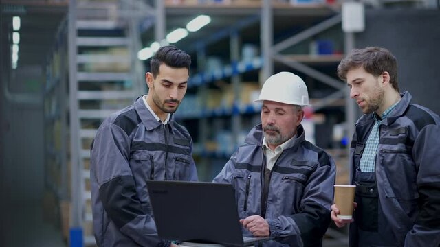 Senior expert man in hard hat showing logistics plan on laptop to employees talking pointing up at stock racks. Three confident Caucasian and Middle Eastern men discussing shipping in warehouse