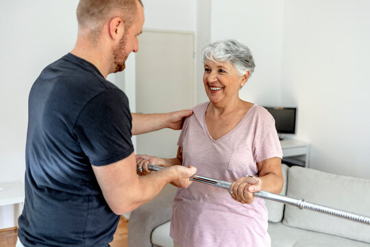 Happy Beautiful Senior Woman Smiling And Holding Easy Bar While Exercising With Personal Trainer. An Older Woman Lifting Weights In Rehabilitation Center. Elderly Lady Is Exercising With Man At Home