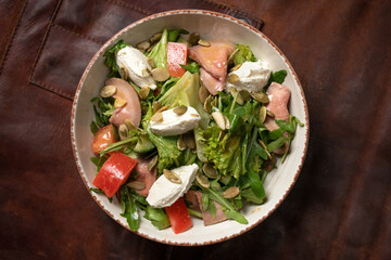 Salmon fresh vegetable salad with mozzarella cheese stands on a table covered with a leather rag and a decorative detail of a simple mechanism. KETO diet concept. OMAD concept. Whole30 concept. 