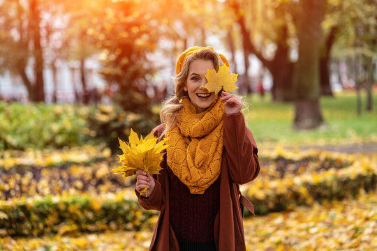 Happy hiding eye woman with a yellowed leaf in yellow knitted beret with autumn leaves in hand and fall yellow garden or park. Beautiful smiling young woman in autumn foliage. 