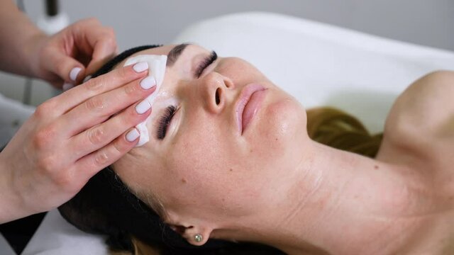 Woman lying with closed eyes on couch in beauty salon, hands of cosmetologist using tissue for removing mask from her face. Closeup skin care procedure. Concept of cosmetology