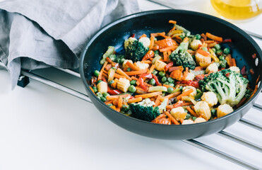 Fried vegetables mix in a black pan on the white table. Healthy food