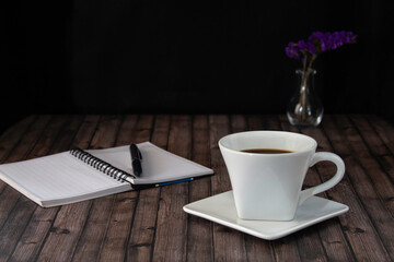 Cup of coffee, notebook and a pen on the wooden table with black background and copy space