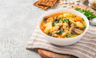 Soup with barley, beans, mushrooms, potatoes, carrots, celery, and herbs. Barley soup with vegetables in a white bowl on a gray concrete background. Vegan food, homemade soup. Free space for text
