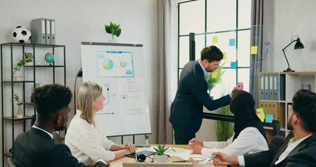 Office work concept where good-looking confident skilled mixed race team sitting at workplace and watching how their successful bearded main manager writing on sticky notes during briefing