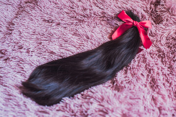 Mechón de pelo cortado anudado con un lazo de raso rojo sobre un fondo de color rosa. Donación de cabello para pelucas para pacientes con cáncer - obrazy, fototapety, plakaty