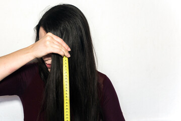 Mujer morena sosteniendo una cinta métrica amarilla para medir la longitud de su cabello largo y negro - obrazy, fototapety, plakaty