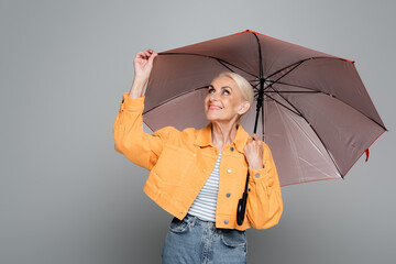 cheerful senior woman in orange jacket touching umbrella isolated on grey