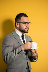 Enjoying his cup of coffee, handsome young bearded business man in eye glasses wearing grey business suit holding cup of coffee standing sideways isolated on yellow background. 