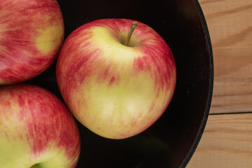 Three juicy apples in a black ceramic plate on a wooden table, top view.