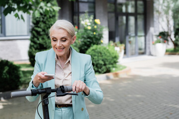 senior businesswoman smiling while messaging on smartphone near electric kick scooter