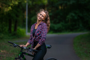 Fototapeta na wymiar Portrait of a smiling young woman in a shirt and leggings on a bicycle standing on an asphalt road in a park on a summer day and looking at the camera, copy space