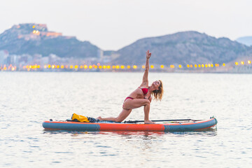 Attractive blonde woman practicing Sup yoga by the sea at sunset with the city lights behind