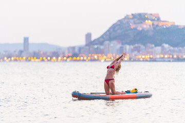Attractive blonde woman practicing Sup yoga by the sea at sunset with the city lights behind