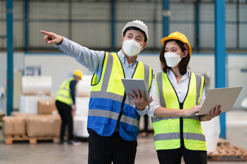 Asian male supervisor engineer in helmet safety wearing hygienic mask to protect coronavirus with female technician holding laptop point to storage location of products in warehouse factory industrial - obrazy, fototapety, plakaty