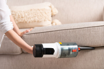 Young woman vacuums sofa with cordless handheld vacuum cleaner. House cleaning. Close-up of vacuum...