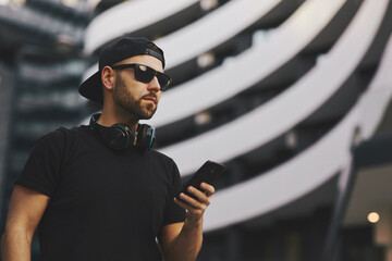 Young man with black hat and headphones holding smartphone in hand in the city