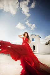 Fashion outdoor photo of gorgeous young woman with red hair in airy red dress posing on the streets of Oia, Santorini, Greece. 