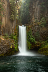 View of a cascading waterfall in Oregon. Toketee Falls is surrounded by green moss, trees, and basalt columns. The waterfall appears to be in a jungle.
