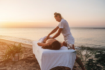 A young masseuse in white clothes makes a massage on the seashore at dawn. Massage table on the...