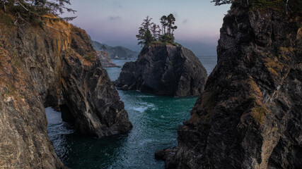 View of cliffs on the coast of Oregon during sunset. Ocean water, rocks, trees, and an arch can be seen in the Samuel H. Boardman Scenic Corridor.