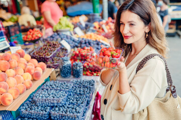 Middle age stylish pregnant woman enjoy shoping, shoosing berries, holding shopping net bag in eco friendly farmer market. Low waste, plastic free lifestyle concept. Eco reusable shopper outdoors.