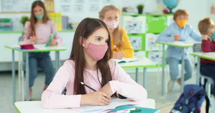 Little pretty Caucasian smart girl junior student in mask sitting at desk in classroom learning at lesson and listening to teacher. Many children studying at school, covid pandemic, Close up portrait