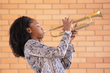 Young afro american woman playing the trumpet with her eyes closed on a brick wall background - obrazy, fototapety, plakaty