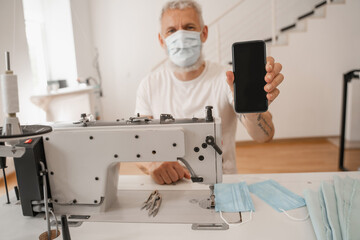 Smartphone in hand of blurred tailor near sewing machine and medical masks
