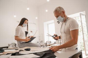 Obraz premium Designer in medical mask holding scissors near cloth and asian colleague