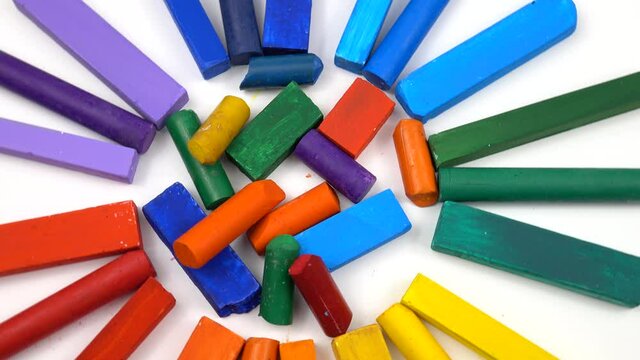Sticks of multicolored chalks in a palette of the rainbow colors lay in a circle with the pieces of the wax crayons in a center on a white background