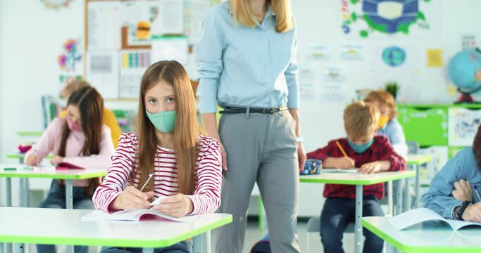 Caucasian teen pretty school student girl solving task with cheat sheet at exam test in class while teacher walking behind. Covid pandemic education, elementary school, children studying