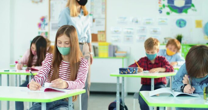Kids at classroom writing examination test. Caucasian teen schoolgirl in medical mask cheating at exam using cheat sheet while female teacher doesn't see. Post-coronavirus education, pupil concept