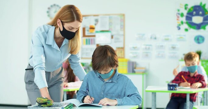 Portrait of Caucasian little junior student writing in copybook studying in classroom listening to tutor Female teacher explaining to smart schoolboy lesson task Elementary education Covid-19 pandemic