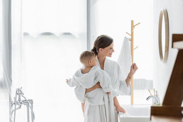 side view of cheerful mother holding in arms toddler son and looking at mirror in bathroom