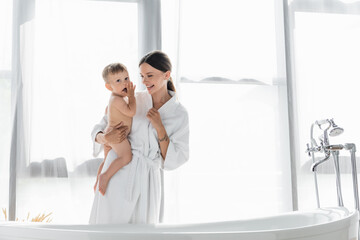 cheerful mother in bathrobe holding in arms naked toddler son near bathtub