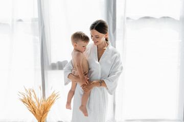 cheerful mother in bathrobe holding in arms naked toddler son