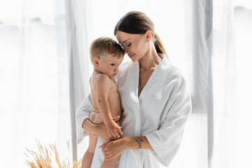 caring mother in bathrobe holding in arms naked toddler son